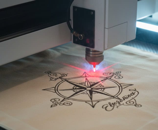 A close-up shot of a CO2 laser cleanly engraving a detailed logo onto a canvas tote bag.